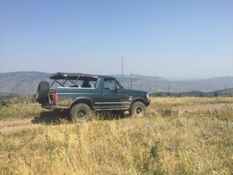 1994 Ford Bronco - A dark green Ford Bronco with a soft top is parked on a dirt road in a dry, grassy field with mountains in the background. The vehicle has…
