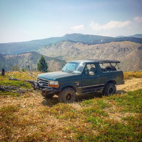 1994 Ford Bronco - A dark green Ford Bronco is parked on a dirt road with mountains in the background. The vehicle has an aftermarket bumper and a soft top.