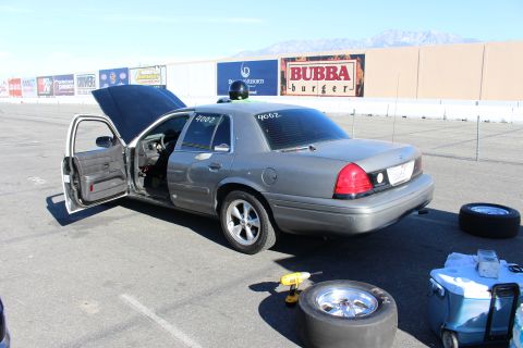 2003 Ford Crown Victoria - gray Ford Crown Victoria sedan sits with its doors open in a paved lot, drag slick tire and cooler nearby, mountains in the distance.