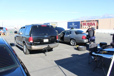 2003 Ford Crown Victoria - black Ford Explorer SUV parks beside a gray Crown Victoria in a lot, a man standing near a fold-out work table with mountains behind.