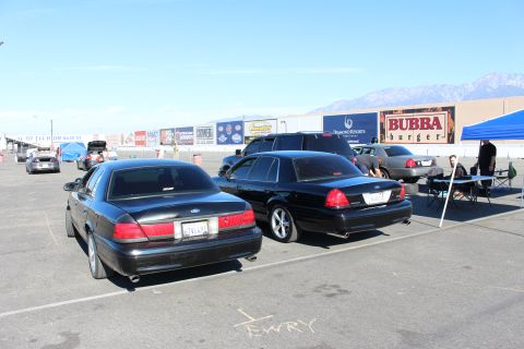 2003 Ford Crown Victoria - Two black Ford and Mercury sedans park nose-to-tail in a lot near a blue canopy tent, mountains and event banners in the background.