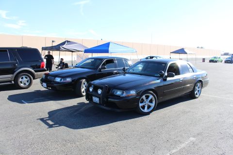 2003 Ford Crown Victoria - black Mercury Grand Marquis and a black Ford Crown Victoria park beside a dark SUV under blue canopy tents in a sunny lot.