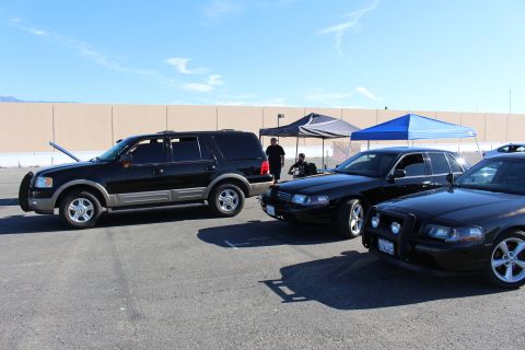 2003 Ford Crown Victoria - black Ford Expedition SUV with hood up parks beside black Ford sedans under canopy tents in a paved lot with a long wall behind.