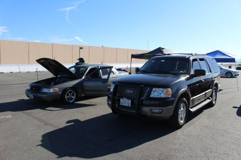 2003 Ford Crown Victoria - black Ford Expedition SUV with a chrome brush guard parks beside a gray Crown Victoria sedan that has its hood and doors open.