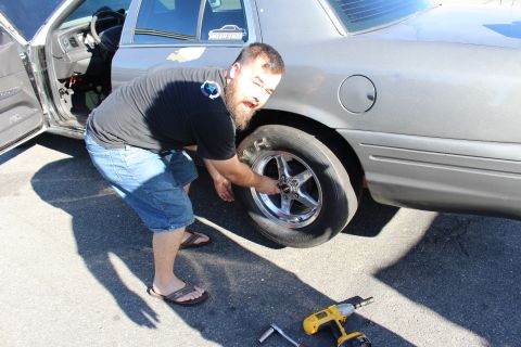 2003 Ford Crown Victoria - bearded man in a dark shirt and denim shorts crouches to mount a chrome drag wheel onto a gray sedan, a cordless drill on the pavement.