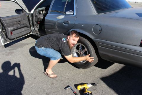 2003 Ford Crown Victoria - bearded man in denim shorts hoists a chrome wheel and tire up to the rear of a gray sedan, hand tools laid out on the pavement.
