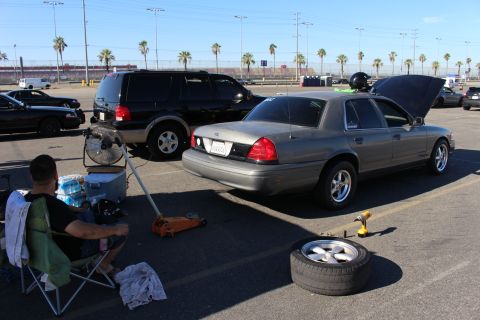 2003 Ford Crown Victoria - gray Ford Crown Victoria sits on a floor jack with hood open beside a black SUV, a spare wheel and tools on the ground, palm trees behind.