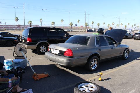 2003 Ford Crown Victoria - gray sedan with open hood and trunk sits on a floor jack next to a black SUV at a paved lot lined with palm trees under blue sky.