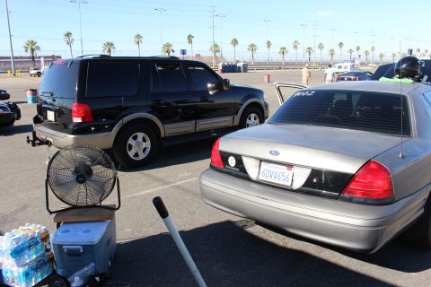 2003 Ford Crown Victoria - black Ford Expedition parks beside a gray Ford Crown Victoria with a standing fan, cooler and water bottles staged at a dragway lot.