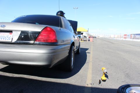 2003 Ford Crown Victoria - Low rear-quarter view of a gray Ford Crown Victoria along a dragway lane, a yellow cordless drill resting on the pavement beside it.