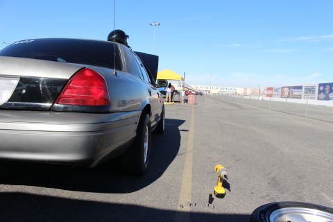 2003 Ford Crown Victoria - Low-angle rear view of a gray sedan parked along a dragway lane, a yellow canopy tent and event banners in the sunlit distance.