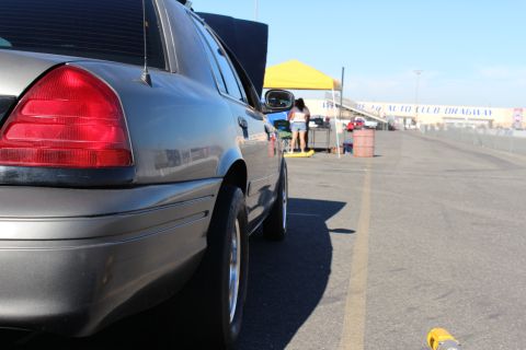 2003 Ford Crown Victoria - Ground-level rear-quarter view of a gray sedan beside a dragway lane, with a yellow tent and Auto Club Dragway wall banner in the distance.