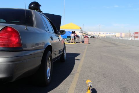 2003 Ford Crown Victoria - Low rear view of a gray sedan with open trunk along a dragway lane, people under a yellow tent and a dragway wall banner beyond.