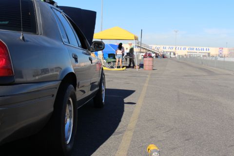 2003 Ford Crown Victoria - Side profile of a gray sedan parked along the Auto Club Dragway lane, people and a yellow canopy tent in the sunlit distance.