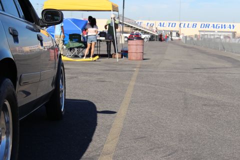 2003 Ford Crown Victoria - Low side view down a dark sedan parked along the Auto Club Dragway lane, with blue and yellow canopy tents and people in the distance.