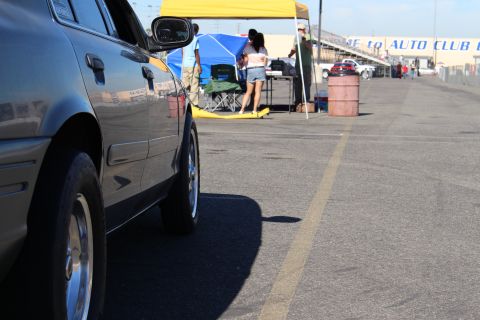 2003 Ford Crown Victoria - Low-angle side view of a gray sedan's front fender and wheel along the Auto Club Dragway lane, with canopy tents and people beyond.
