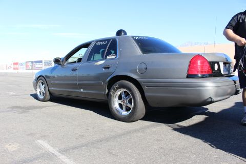 2003 Ford Crown Victoria - Rear-quarter view of a gray Ford Crown Victoria on chrome drag wheels and slick tires in a paved lot, a person standing at right.
