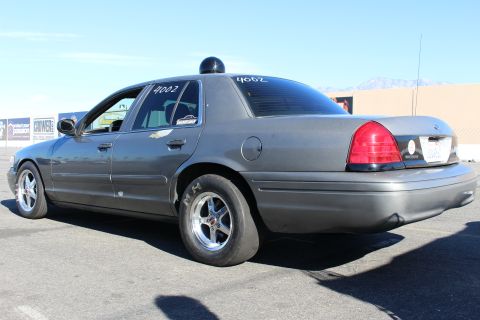 2003 Ford Crown Victoria - same gray Crown Victoria sedan photographed from the rear left, showing chrome drag wheels and a window number under clear skies.
