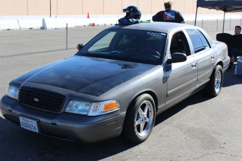 2003 Ford Crown Victoria - gray Ford Crown Victoria with its hood slightly raised and chrome drag wheels, people in helmets standing nearby at a track.