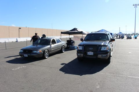 2003 Ford Crown Victoria - gray Ford sedan parked next to a black Ford Expedition SUV on a wide asphalt lot with tents and other vehicles around.