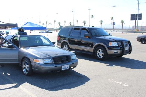 2003 Ford Crown Victoria - gray Crown Victoria with its door open and a black Ford Expedition sit on a lot lined with palm trees and a freeway beyond.
