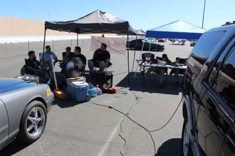 2003 Ford Crown Victoria - group of people relax in folding chairs under a pop-up canopy with a cooler and fan, flanked by a gray car and a black SUV.