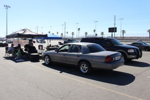 2003 Ford Crown Victoria - gray Ford Crown Victoria sits beside a dark SUV while people lounge under tents at a sunny motorsport venue with palm trees.