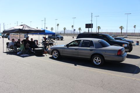 2003 Ford Crown Victoria - gray Ford Crown Victoria sits on a wide lot as people rest under a canopy, with palm trees and a grandstand in the distance.