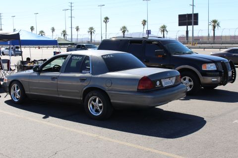 2003 Ford Crown Victoria - gray Ford Crown Victoria seen from the rear left, parked among dark SUVs on a sunny lot lined with palm trees.