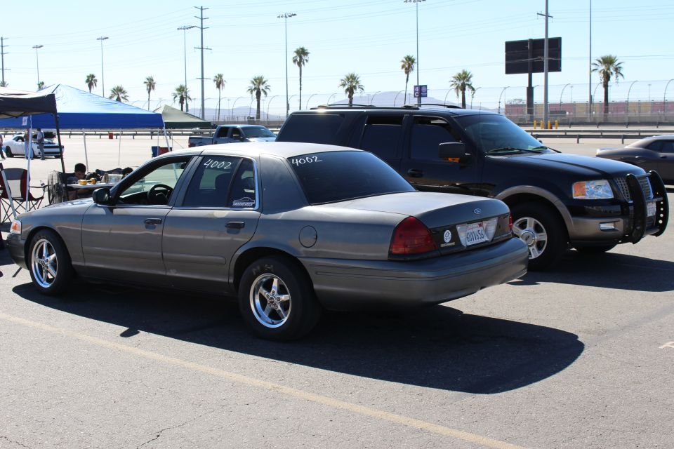 Rear left view of a gray Ford Crown Victoria beside dark SUVs in a parking lot
