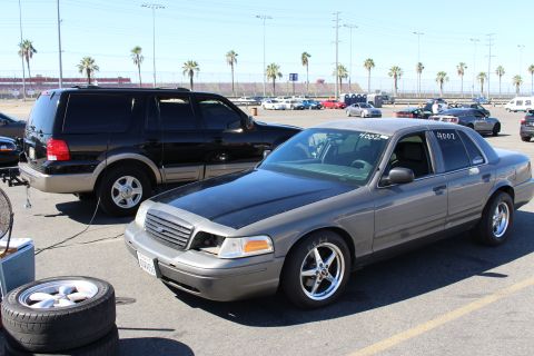 2003 Ford Crown Victoria - gray Ford Crown Victoria with chrome drag wheels sits beside a loose spare wheel, a black SUV and palm trees behind.