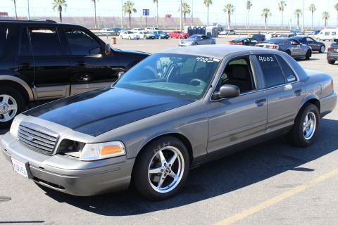 2003 Ford Crown Victoria - gray Ford Crown Victoria with a matte-black hood and chrome drag wheels parks in a lot full of cars under palm trees.