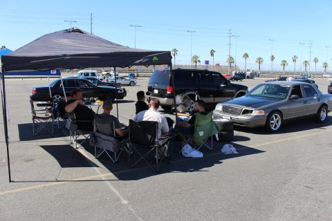 2003 Ford Crown Victoria - group sits in folding chairs under a dark canopy as a gray Ford sedan and SUVs line a sunny lot with palm trees behind.