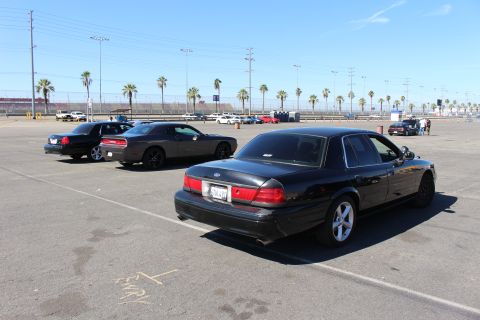 2003 Ford Crown Victoria - black sedan with a Ford-style trunk emblem sits in the foreground among other dark cars on a sunny palm-lined lot.
