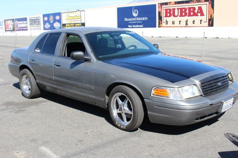 2003 Ford Crown Victoria - gray Ford Crown Victoria with a matte-black hood and chrome drag wheels sits on a lot before billboards including Bubba Burger.