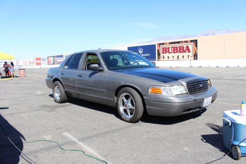 2003 Ford Crown Victoria - gray Ford Crown Victoria with a black hood and chrome drag wheels parks beside a cooler, with billboards on the building behind.