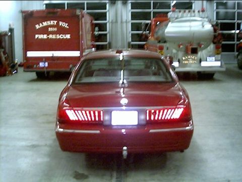 2000 Mercury Grand Marquis - Rear view of a red 2000 Mercury Grand Marquis parked inside a fire station with two fire trucks behind it.