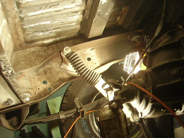 Underside of a vehicle showing frame, leaf spring, shock absorber, and drivetrain components.