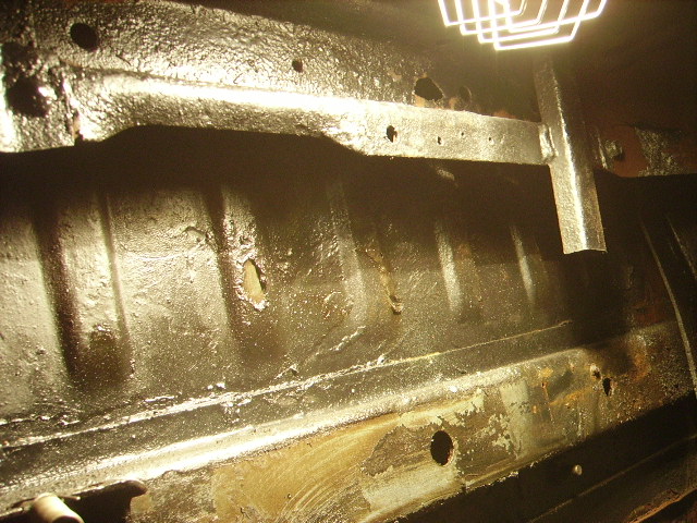 Underside of a vehicle chassis with rust and holes, showing a welded bracket and overhead light.
