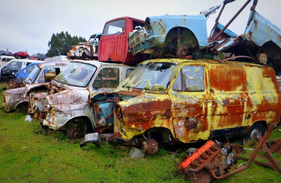 A junkyard scene with multiple rusted vehicles piled up, including a yellow van and a red truck.