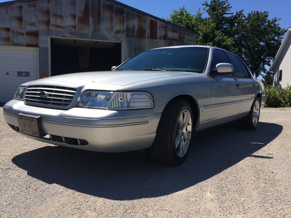 Silver Ford Crown Victoria parked in front of a rustic garage. The car has aftermarket wheels and a license plate