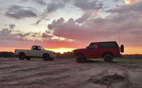 1995 Ford Bronco - A red Ford Bronco and a white Ford Ranger pickup truck are parked on a dirt track during sunset.