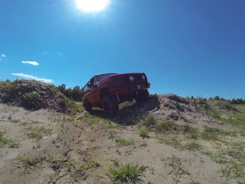 1995 Ford Bronco - A red 1995 Ford Bronco is shown from a low angle, driving up a sandy hill under a bright blue sky with the sun shining.