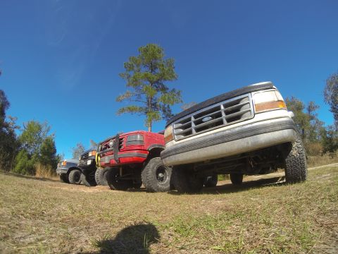 1995 Ford Bronco - A low-angle, wide-angle shot of four off-road modified trucks parked on a grassy field under a clear blue sky. The closest truck is a white…