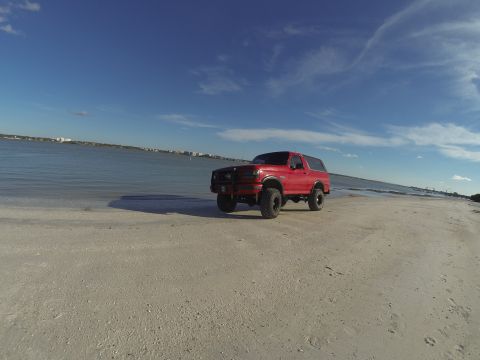 1995 Ford Bronco - A lifted red Ford Bronco is parked on a sandy beach next to the ocean under a bright blue sky with scattered clouds.