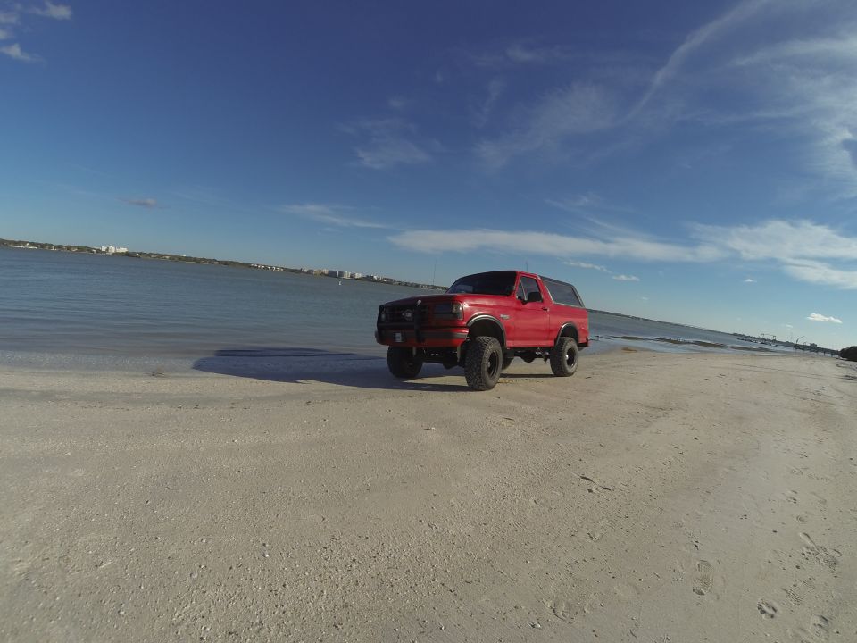 Red lifted Ford Bronco on a sandy beach by the ocean.