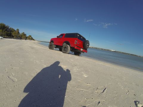 1995 Ford Bronco - A red, lifted Ford Bronco is parked on a sandy beach next to the ocean under a clear blue sky. The vehicle is viewed from a low angle, capt…