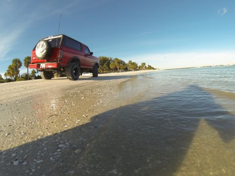 1995 Ford Bronco - A red Ford Bronco is parked on a sandy beach next to the ocean. The vehicle is viewed from a low angle, showing its rear and side profile w…