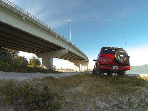 1995 Ford Bronco - A red 1995 Ford Bronco is parked on a sandy area near a bridge and the ocean. The spare tire on the back has a "Ford Bronco" logo.