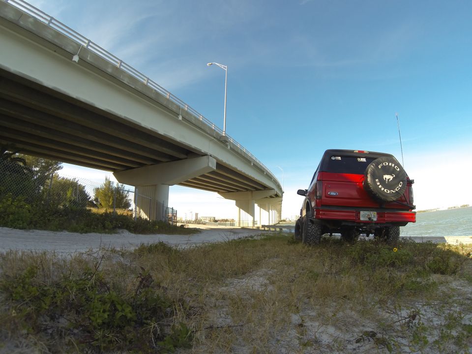 Red 1995 Ford Bronco parked under a bridge with the ocean in the background.